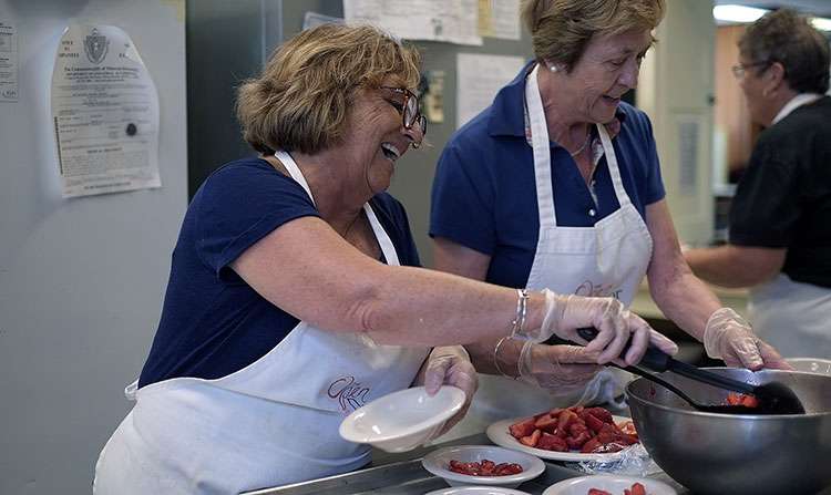 Two women in aprons smile while serving a community meal.