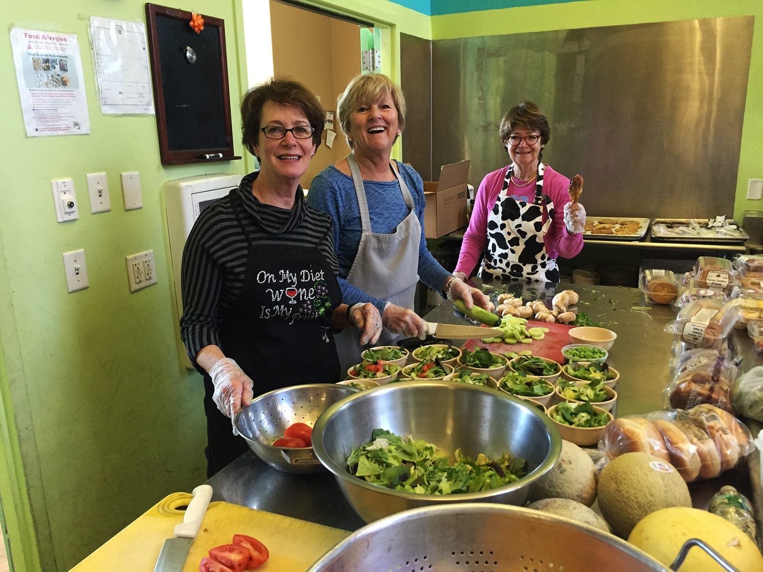 A client smiles at the camera while waiting for lunch at a Lifebridge community meal. Salads and desserts are individually plated for pick up while two kitchen staff prepare a grilled sandwich for him in the background