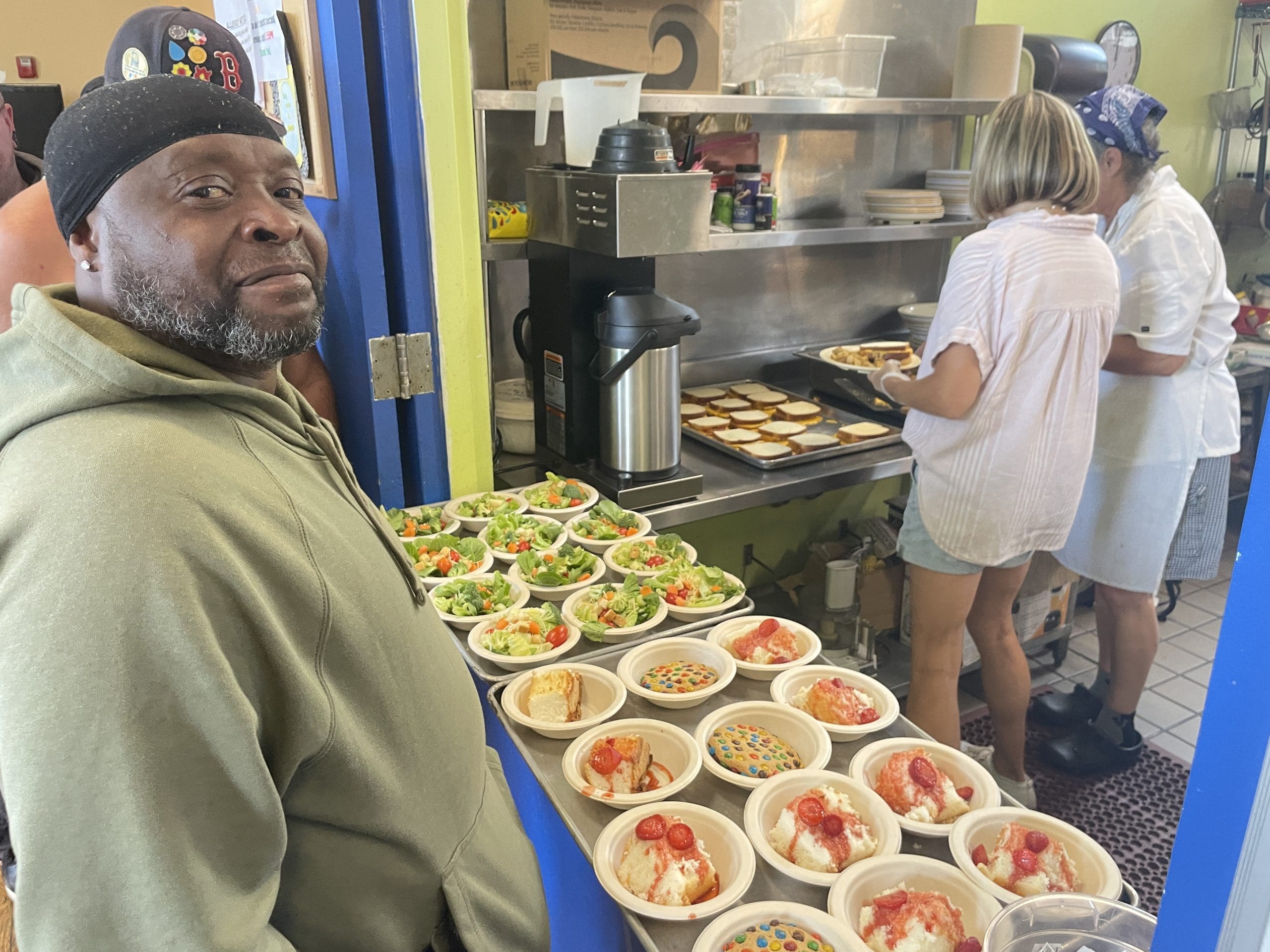 A client smiles at the camera while waiting for lunch at a Lifebridge community meal. Salads and desserts are individually plated for pick up while two kitchen staff prepare a grilled sandwich for him in the background
