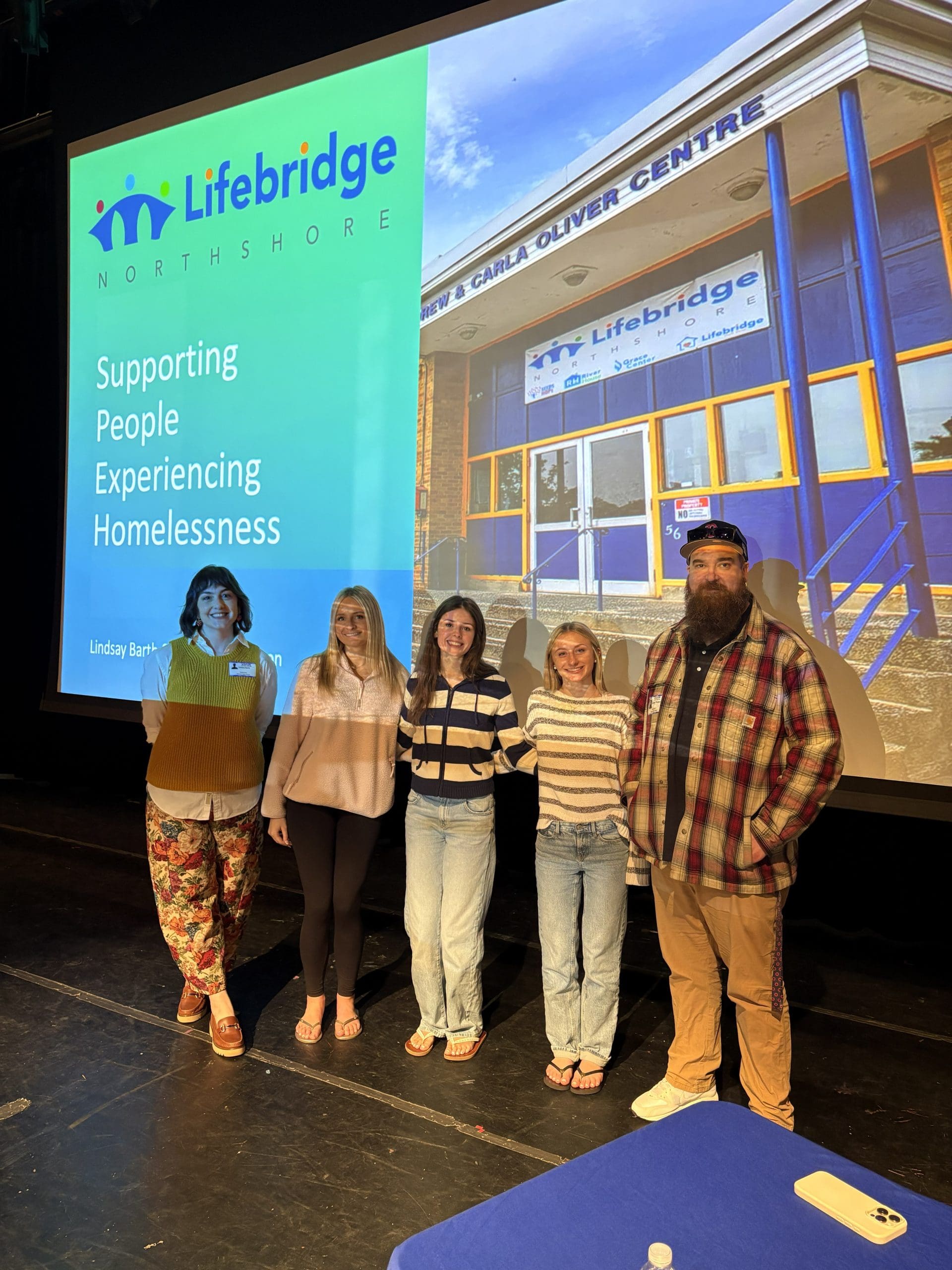 Lindsay and George pose with Danvers High School Students on a stage in front of a screen that says "Lifebridge: Supporting People Experiencing Homelessness"